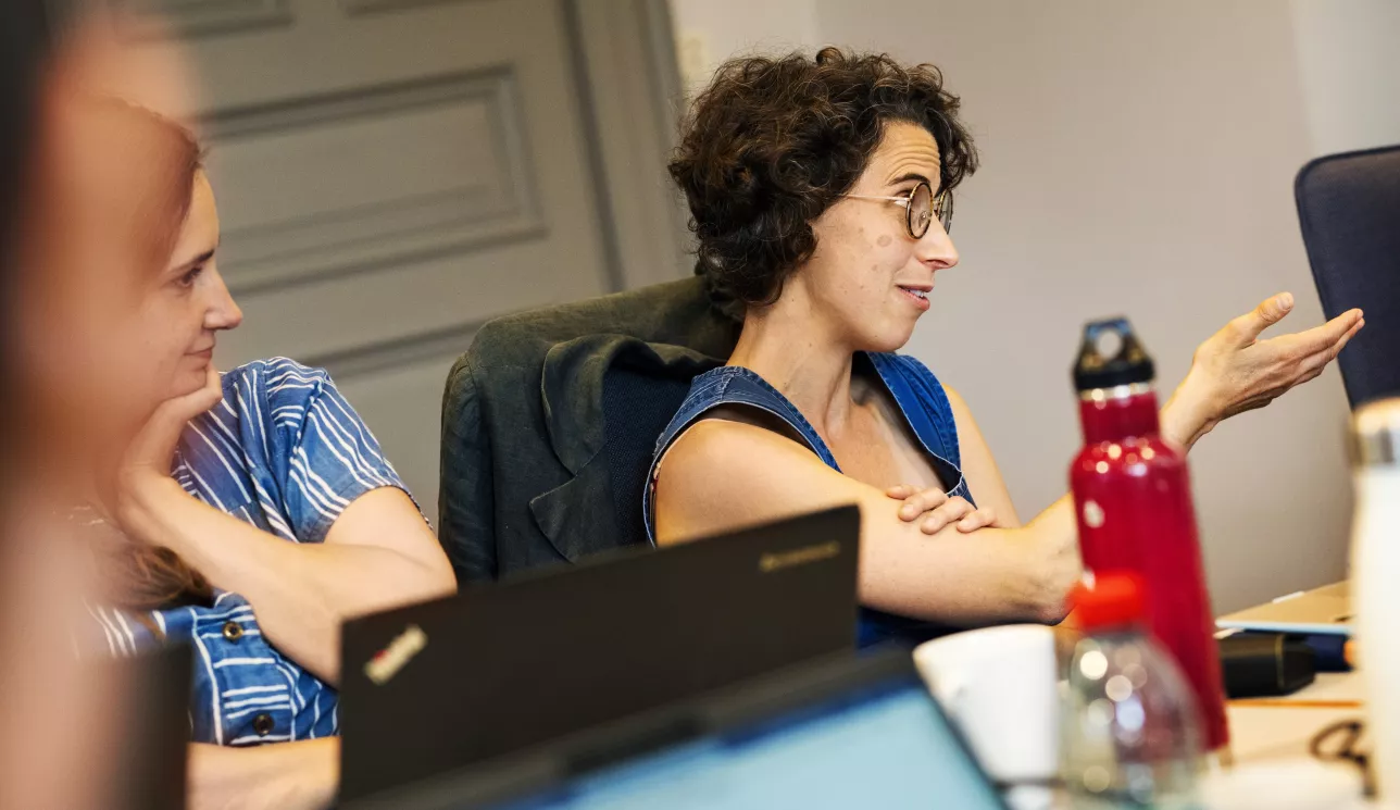 Young woman at a table filled with laptops and water bottles gestures while talking. Photo.