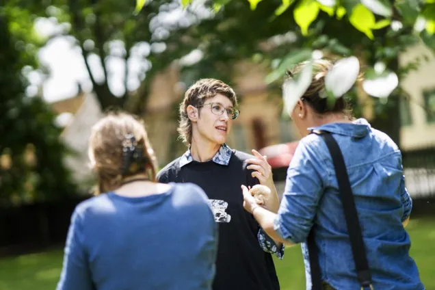 Ung kvinna samtalar med två personer med ryggen åt kameran utomhus. Foto.