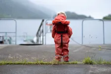 Ett ensamt barn i flytväst står med ryggen mot kameran och tittar ut över grått hav. Foto.